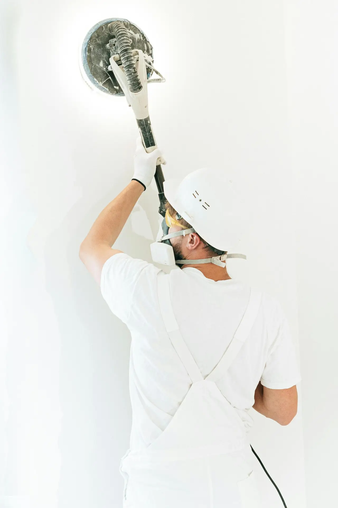 Construction worker smoothing a wall with a power sander, wearing safety gear and white overalls.